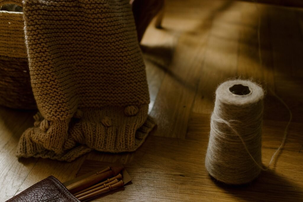From above of handmade woolen blanket placed in wicker basket near various wooden knitting needles located on parquet floor with spool of thread